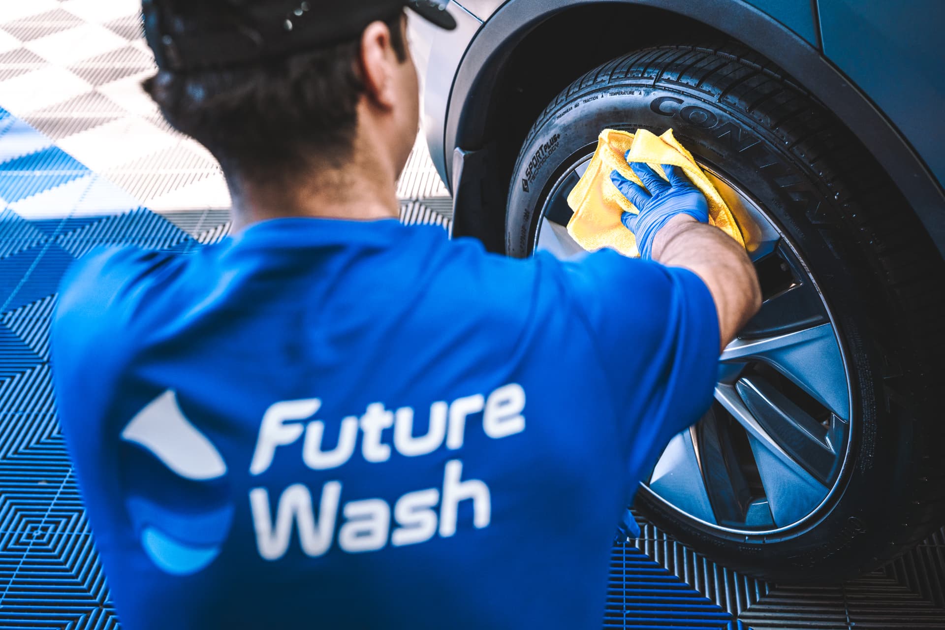 Future Wash technician in a branded blue shirt wiping down a wheel with a microfiber towel in the retail bay