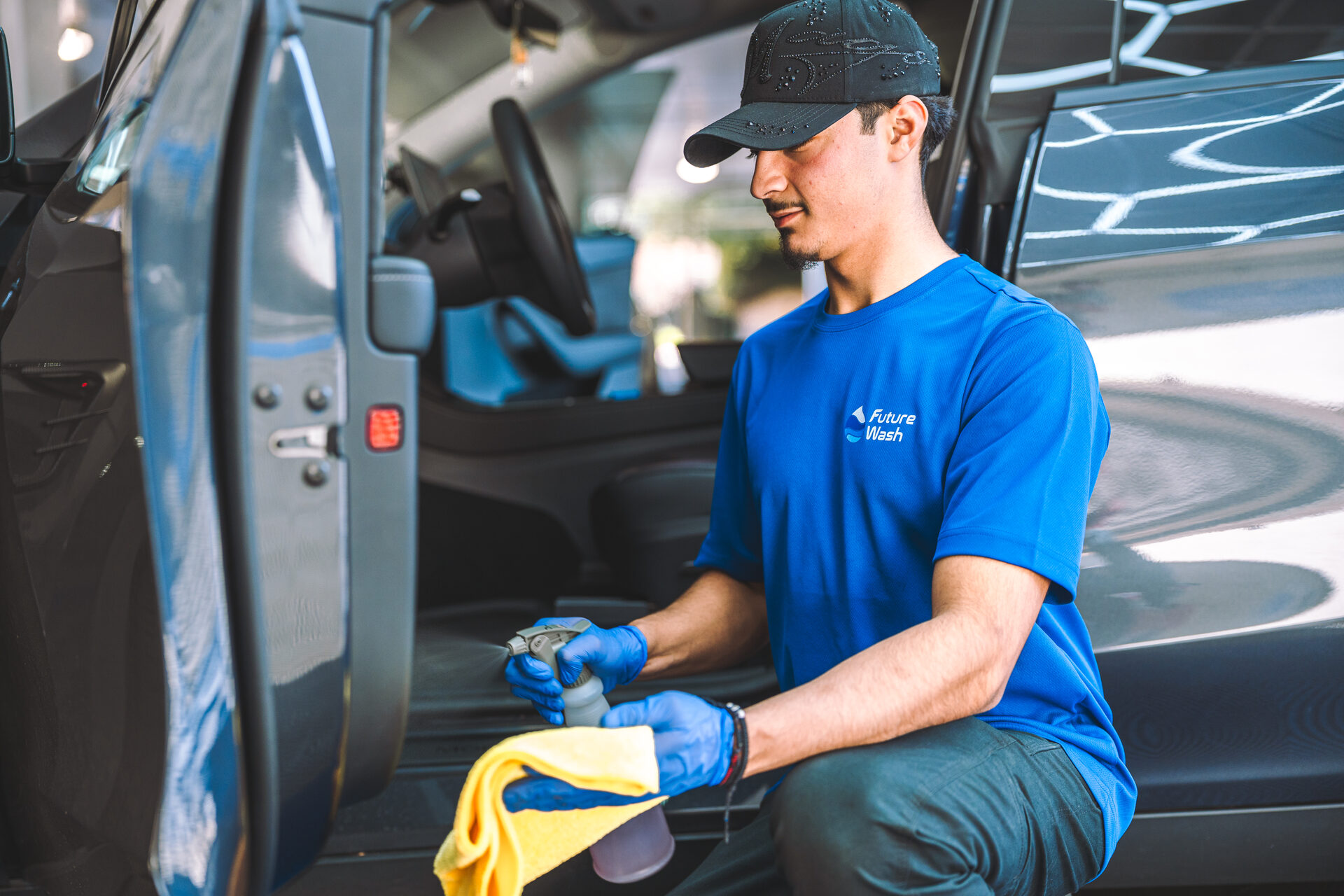 Future Wash technician kneeling in the doorway of a dark SUV with a spray bottle and microfiber cloth in a parking garage